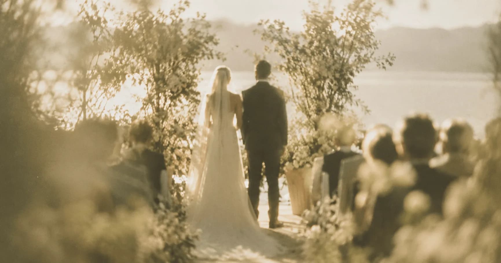 Escena onírica de una boda vista en la distancia: una pareja de novios de espaldas camina hacia un lago bajo una luz dorada